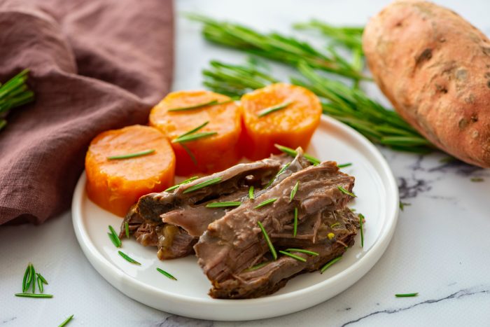 white plate with cooked roast beef with sweet potatoes, garnished with rosemary