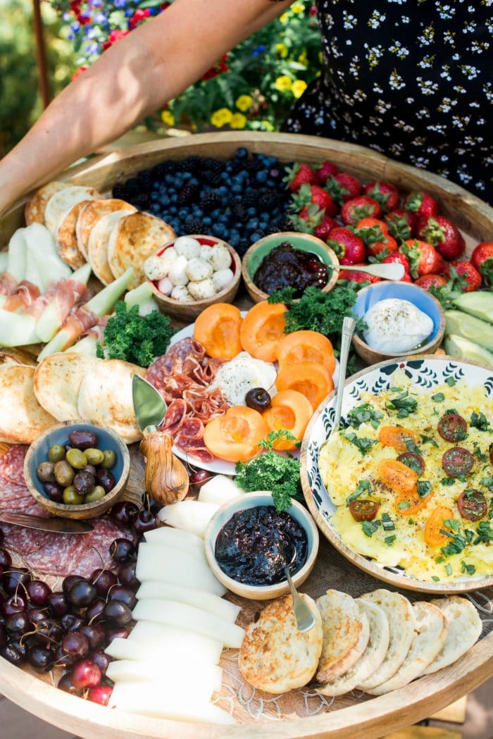 a round board filled with beautiful fruit, English muffins, a bowl of scrambleld eggs, meat, and cheese