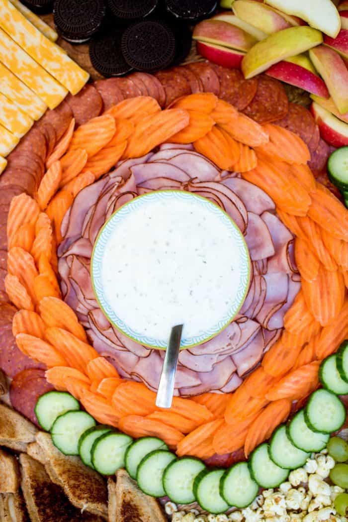 Bowl of Ranch dressing surrounded by ham slices, carrot slices, cucumber slices, and pepperoni