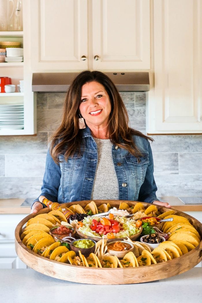 lady holding a large round platter surrounded with tacos and toppings