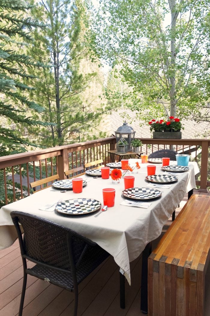 long table on deck outside with trees with table cloth and plates and cups