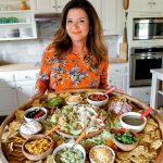 In kitchen holding a big wood board filled with bowls of chicken, rice, and taco toppings. Surrounded by a lot of chips.