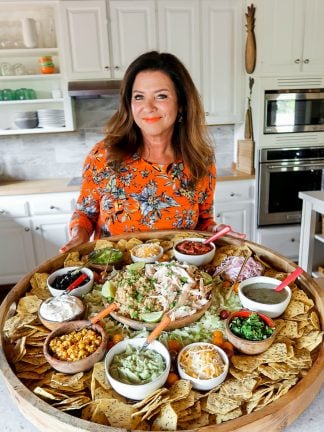 In kitchen holding a big wood board filled with bowls of chicken, rice, and taco toppings. Surrounded by a lot of chips.