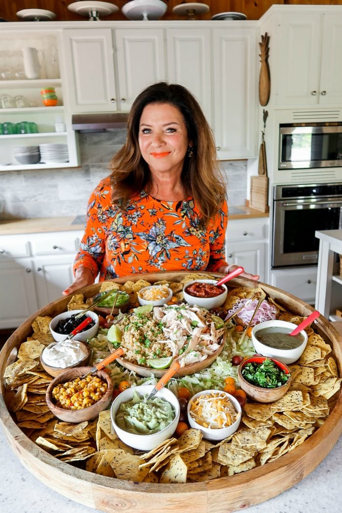 In kitchen holding a big wood board filled with bowls of chicken, rice, and taco toppings. Surrounded by a lot of chips.