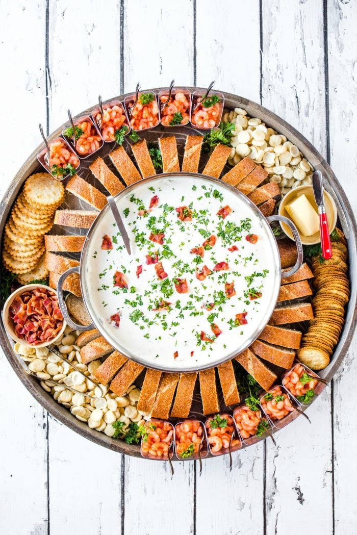 a beautiful dinner board with a pot of clam chowder in the middle, with bread, crackers, and shrimp cups