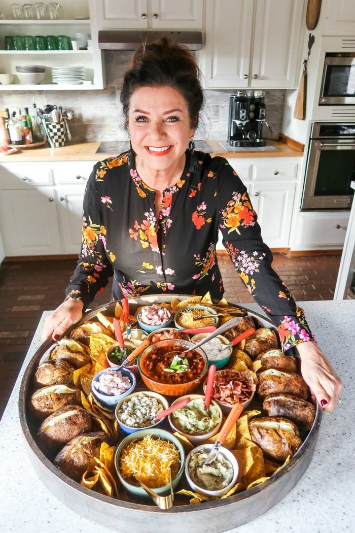 woman holding an epic round wood board with baked potatoes and small bowls of toppings