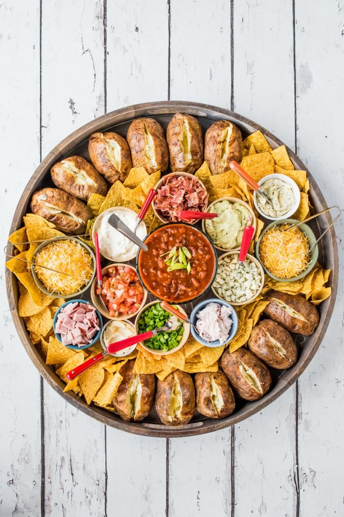 an epic round board filled with plain baked potoatoes and bowls of different toppings