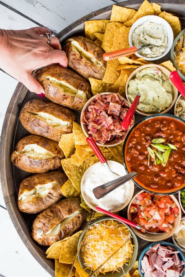 a woman's hand grabbing a baked potato (on a board with a bunch of hot baked potatoes and toppings)