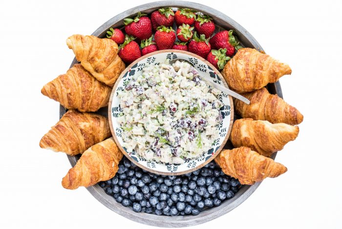 looking down on a wood board with chicken salad sandwich mixture in a separate bowl in the middle, surrounded by croissants, and fresh berries