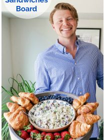 man holding a large wood board filled with chicken salad, croissants, and fresh berries