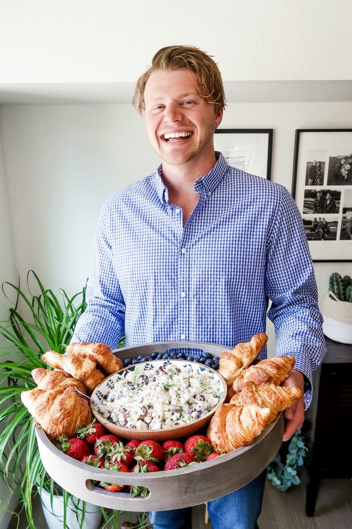 man laughing holding a large wood board filled with chicken salad, croissants, and berries