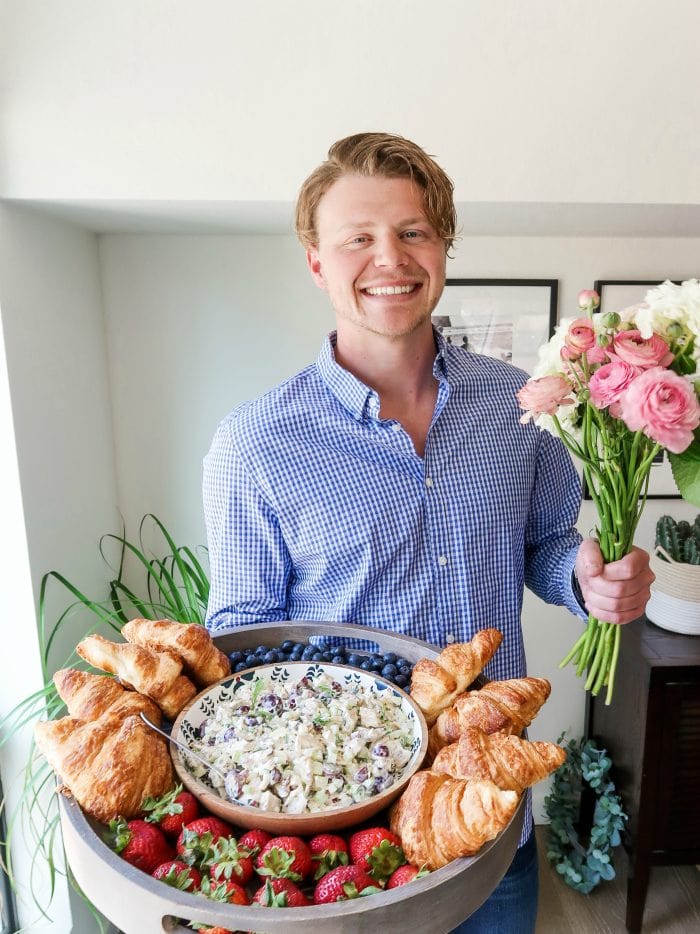 man holding a large wood board filled with chicken salad, croissants, and fresh blueberries and strawberries and fresh flowers
