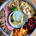a round board with crackers, salami, fruit, nuts, and brie in the center