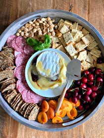 a round board with crackers, salami, fruit, nuts, and brie in the center