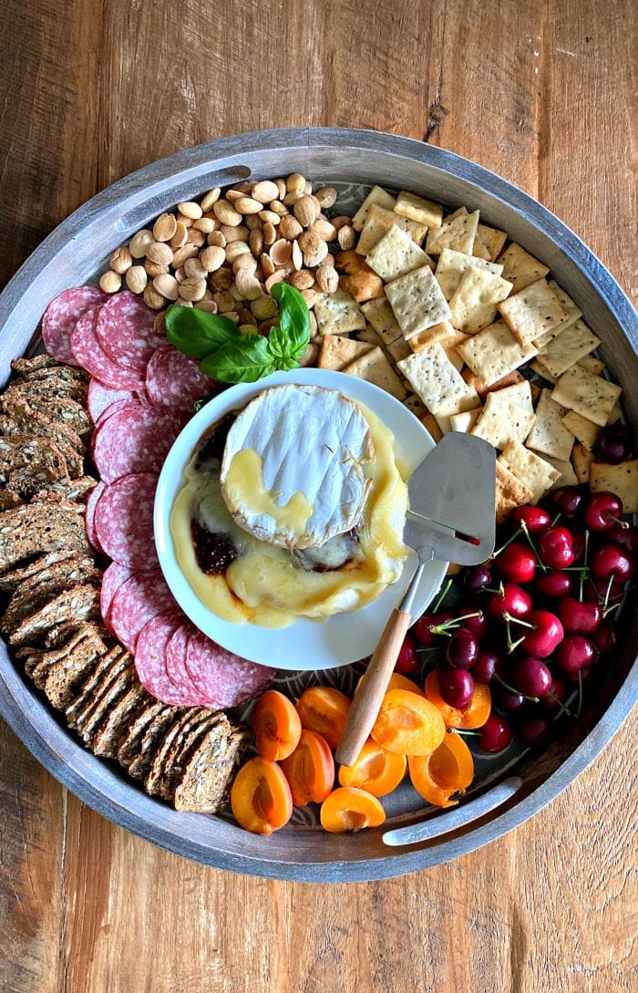 a round board with crackers, salami, fruit, nuts, and brie in the center