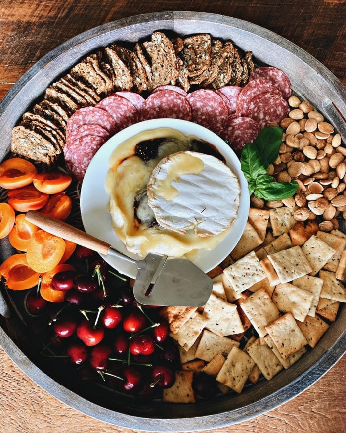 beautiful round board with brie cheese in the middle, surrounded by crackers, fresh fruit, nuts