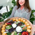 woman holding a round tray of stacked crackers, soft cheese, veggies, and herbs
