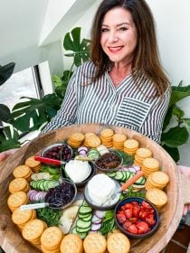 woman holding a round tray of stacked crackers, soft cheese, veggies, and herbs