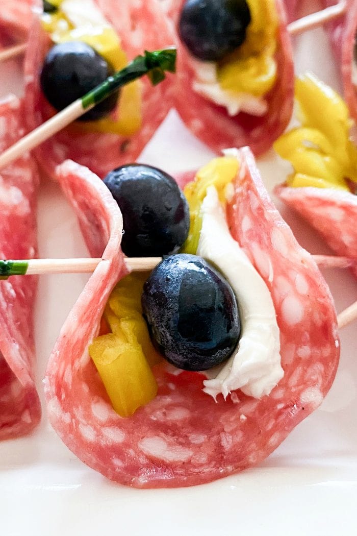 close up of one salami slice with cream cheese spread on top, with 2 blueberries, a piece of pepperoncini, folded together with a toothpick