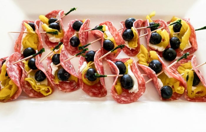 a platter of salami slices with cream cheese spread on top, with 2 blueberries, a piece of pepperoncini, folded together with a toothpick