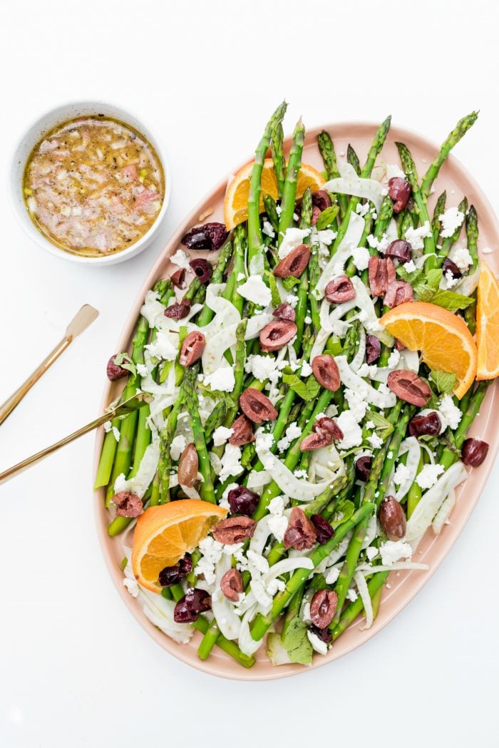 Shaved Fennel Asparagus Salad on an oblong platter wth a small bowl of orange dressing and gold serving tongs