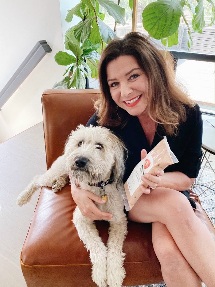 woman holding a bag of dog food in one hand and arm around a Whoodle dog in the other