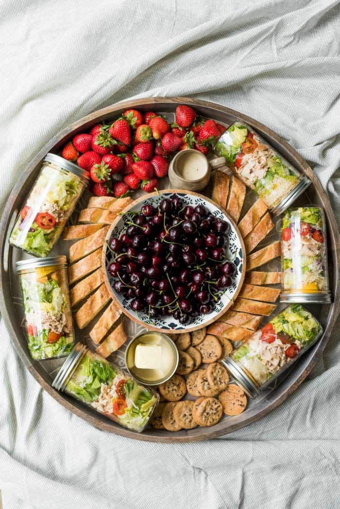 a round board with jars of salad, baguette bread, strawberries, and a bowl of cherries in the middle