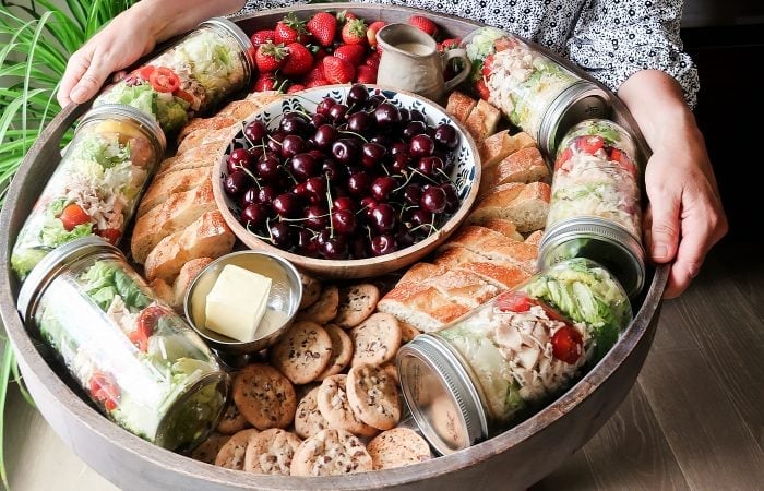a big board of food with cherries in the middle, with jars of salad, bread, and cookie