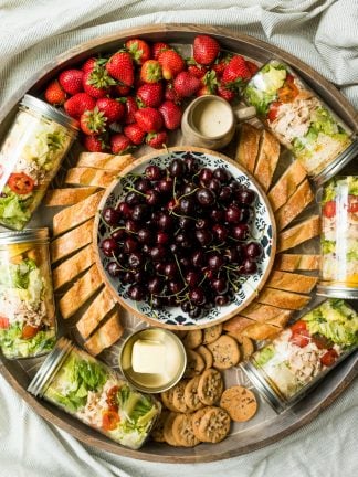 a round board with jars of salad, bread, strawberries, and a bowl of cherries