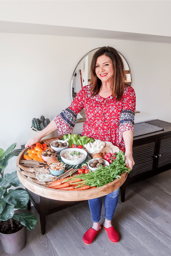 woman holding a very large round board filled with vibrant veggies (cut up) with a bowl in the center of cashew dip, with other Vegan foods (meatballs, pita, etc.)
