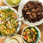 a taco spread with meat, corn salad, salsa, tortillas, and green sauce