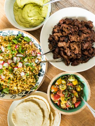 a taco spread with meat, corn salad, salsa, tortillas, and green sauce