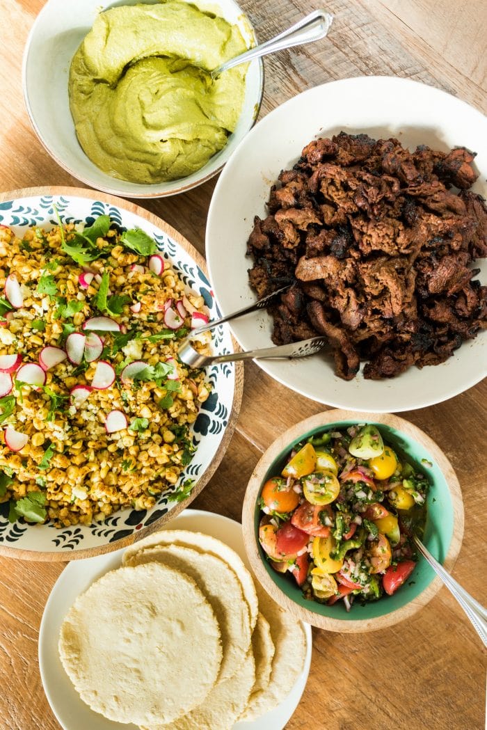 a taco spread with meat, corn salad, salsa, tortillas, and green sauce