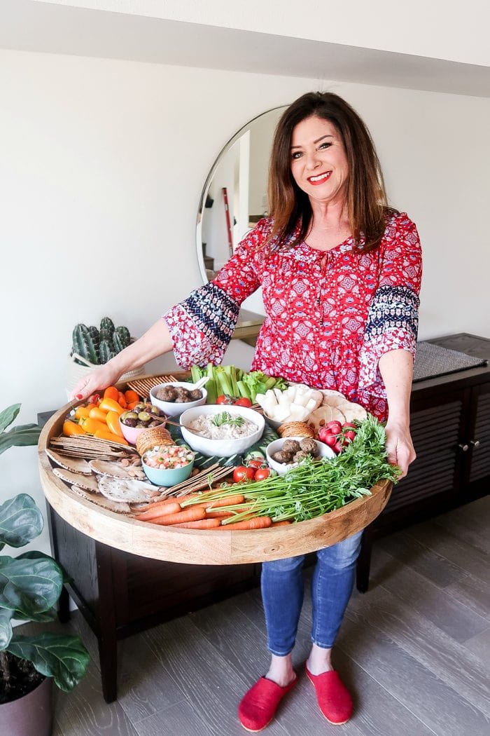 woman in red blouse holding an epic wood board filled with crudite veggies, dips, and more