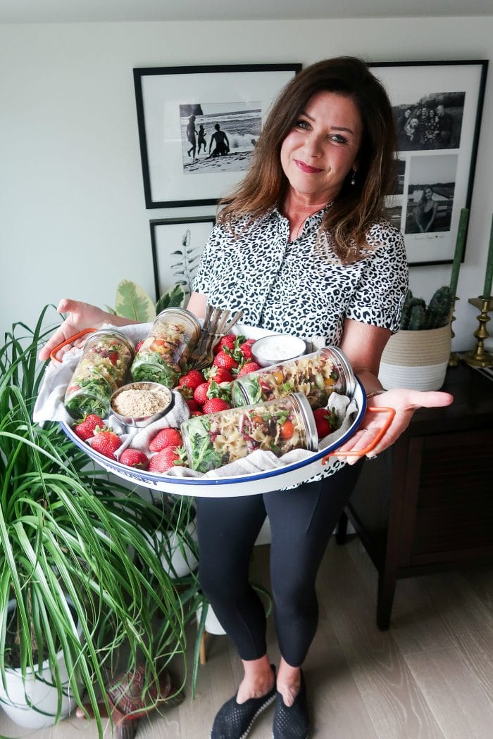 woman in black and white shirt holiding an oval tray with 4 jars of pasta salad, and strawberries