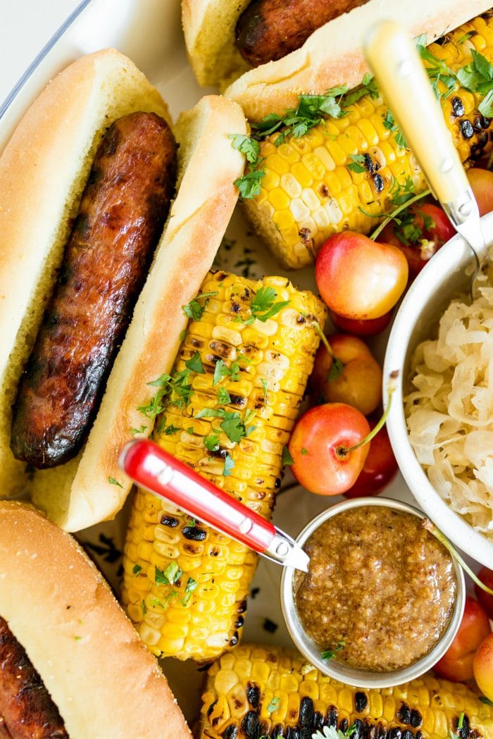 close up grilled brat on bun and grilled corn with mustard in small bowl
