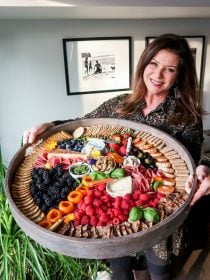 woman holding a large round board with crackers, cheese, meets, and fruit