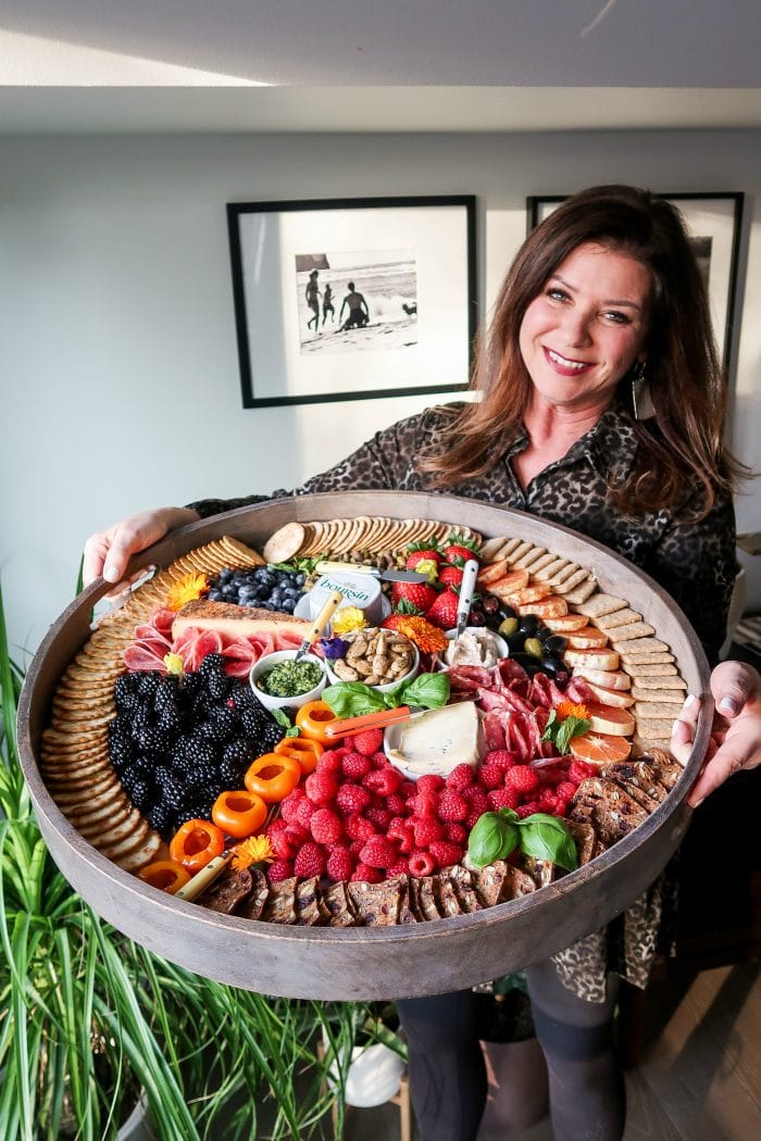 woman holding a large round board with crackers, cheese, meets, and fruit