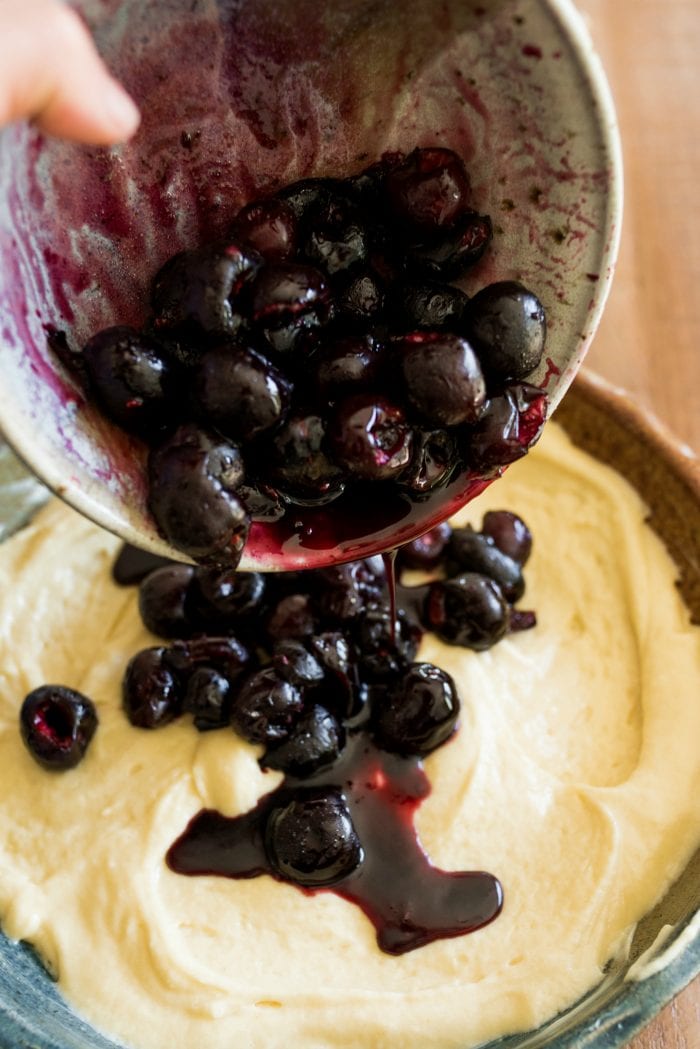 pouring juicy red cherries into buttermilk cake batter in a round pan