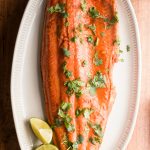 one large salmon filet on a white plate, cooked with lime and garlic, garnished with lime and cilantro