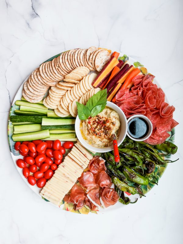 a round tray with cheese, meat, crackers, veggies, and hummus