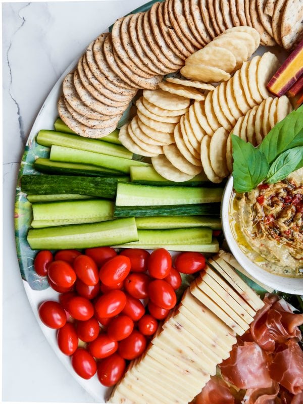 tomatoes, cucumber sticks, and crackers on a cheese board with hummus