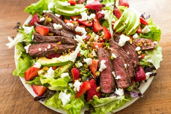 round plate of green salad with steak, avocados, strawberries, and cheese