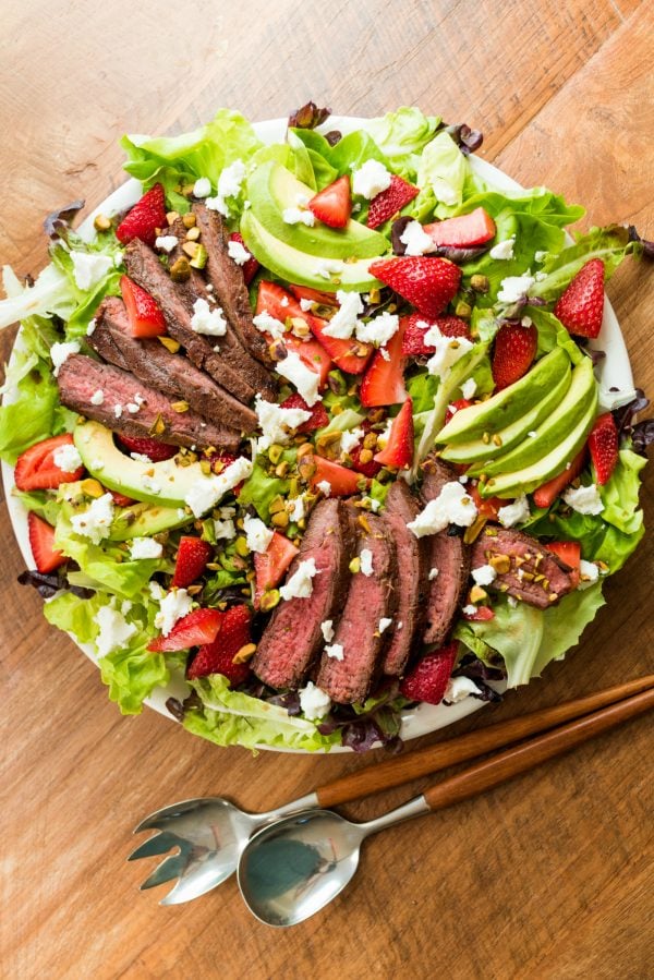 round plate of green salad with streak, avocados, and strawberries