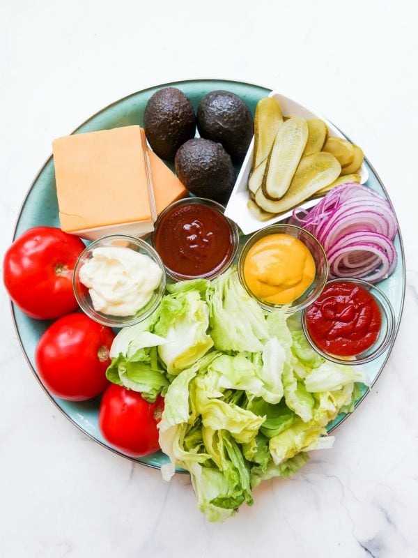 platter of iceberg lettuce, whole tomatoes, cheese, avocados, pickles, red onion, and condiments like mayo, mustard, ketchup, BBQ sauce